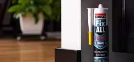 Kartusz kleju-uszczelniacza Soudal Fix All Flexi stojący przy framudze drzwi. Tuba produktu w biało-czarno-czerwonych barwach stoi na betonowej podłodze przy czarnej listwie przypodłogowej i białym słupku. Po lewej stronie widać rozmyty salon z drewnianą podłogą i rośliną doniczkową.