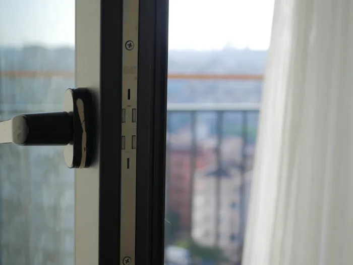 Uchylone drzwi balkonowe, na pierwszym planie klamka, w tle barierki i zamazany widok na one budynki