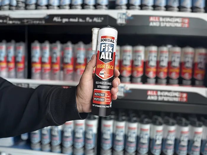 Ręka trzyma kartusz kleju Soudal Fix All High Tack na tle półki z innymi produktami z tej serii