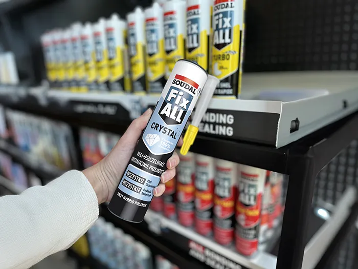 Soudal Fix All Crystal — przezroczysty klej-uszczelniacz hybrydowy trzymany w dłoni na tle regału z produktami Fix All w sklepie