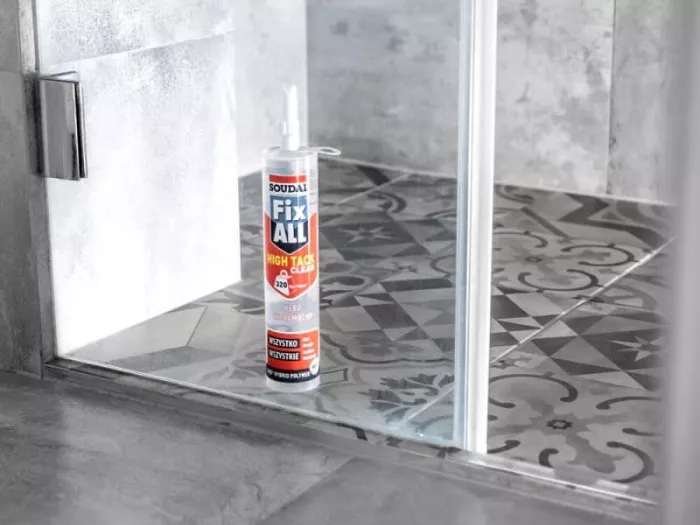 Kartusz kleju Soudal Fix All High Tack Clear stojący na podłodze z ozdobnych szaro-białych płytek ceramicznych, u wejścia do nowoczesnego prysznica ze szklaną kabiną i szarymi kafelkami na ścianie.