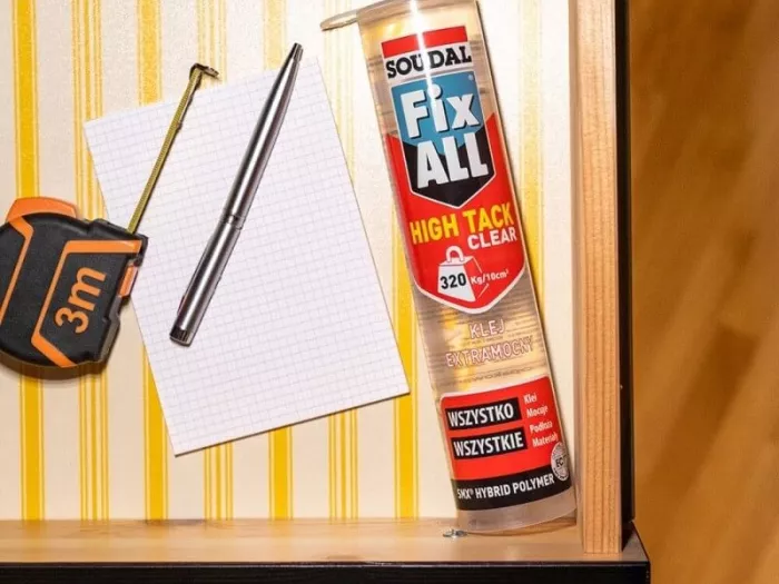 Bezbarwny klej Soudal Fix All High Tack Clear leżący w szufladzie obok metalowego ołówka stolarskiego, kartki papieru w kratkę i zwijanej miarki 3m.