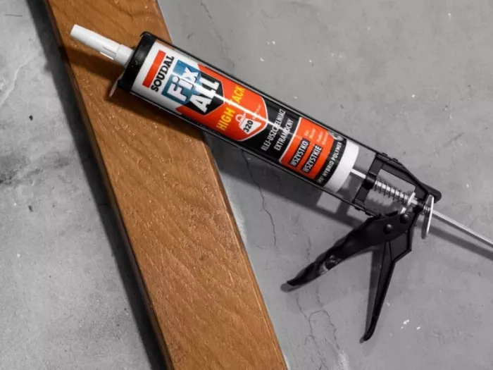 Kartusz czerwonego kleju Soudal Fix All High Tack załadowany do czarnego wyciskacza pistoletowego, leżący obok szerokiej drewnianej deski na betonowej podłodze.