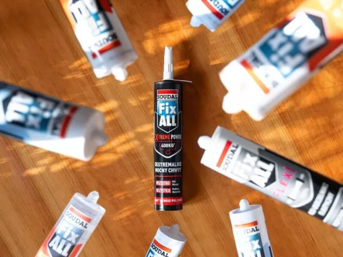 Kartusz kleju Soudal Fix All X-Treme Power umieszczony centralnie na drewnianym blacie, otoczony rozłożonymi promieniście innymi tubami klejów Fix All. Zdjęcie z góry w promieniach słońca.